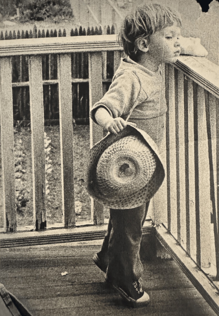 A young child on a porch holding a wide straw hat, photographed by Margery Lee McKeon